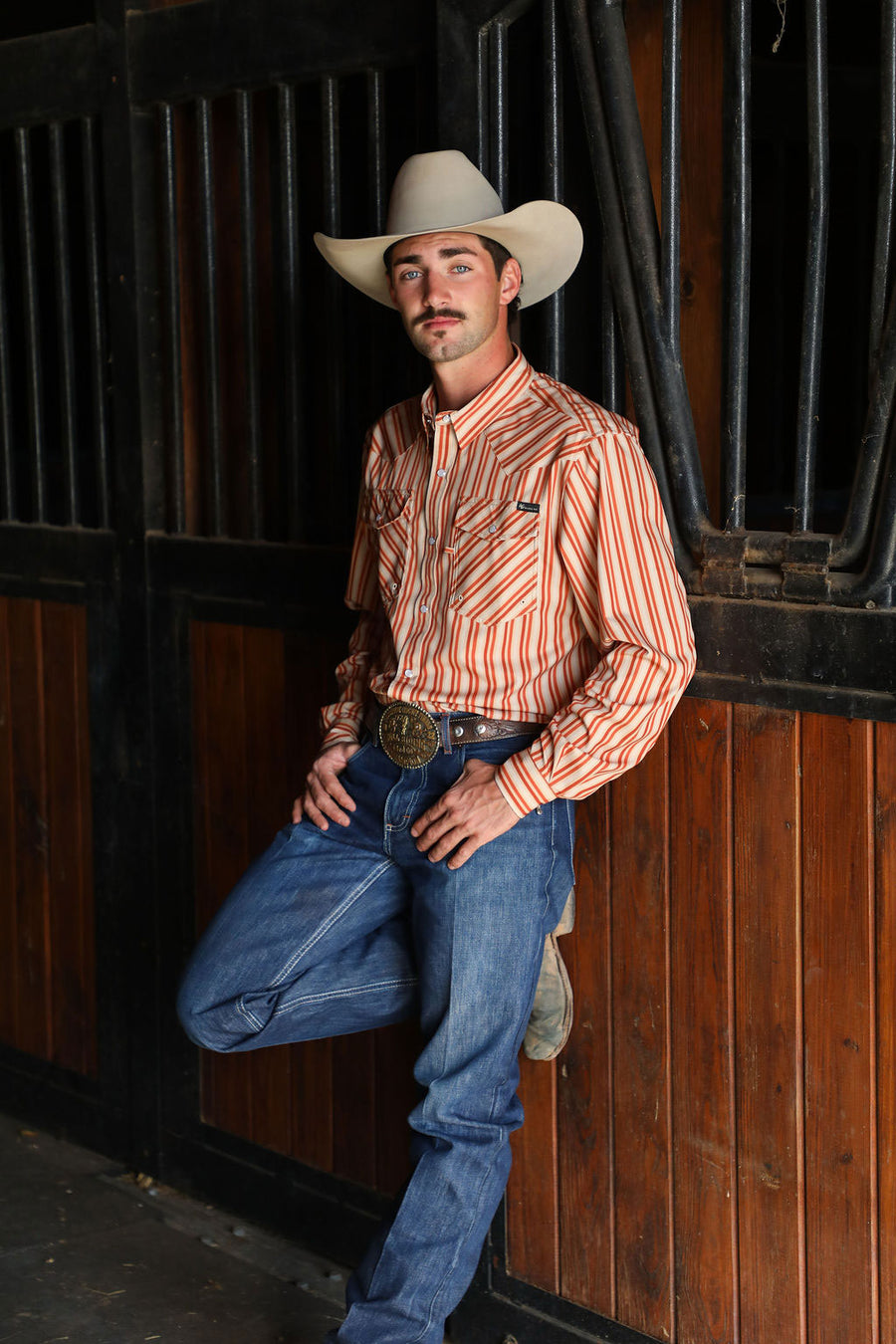 Man wearing a cowboy hat winners way apparel leaning against a horse stall. 