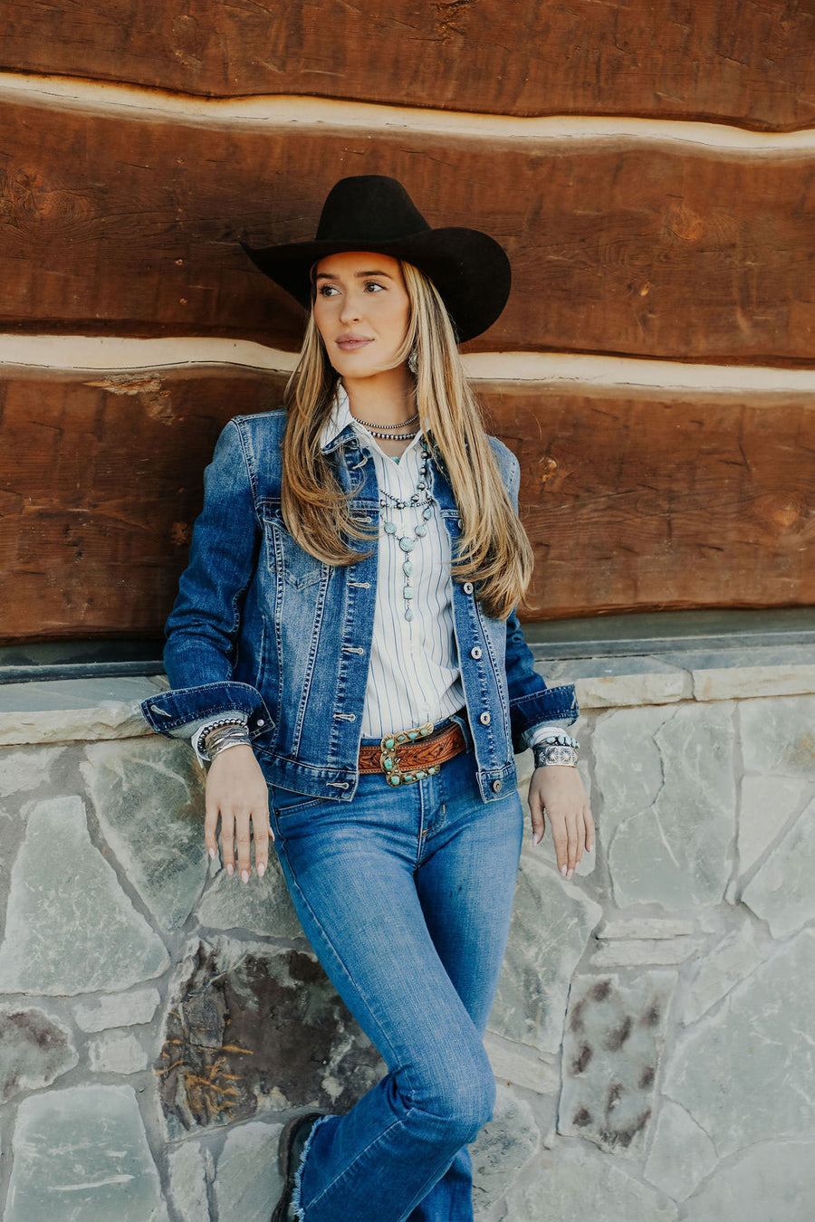 Woman wearing a denim jacket, cowboy hat and winners way apparel leaning against a rustic wooden and stone wall.